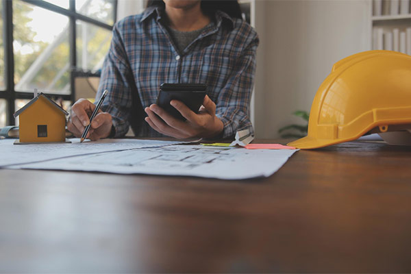 A woman multitasks, using her phone while sitting at a table with construction plans spread out before her.